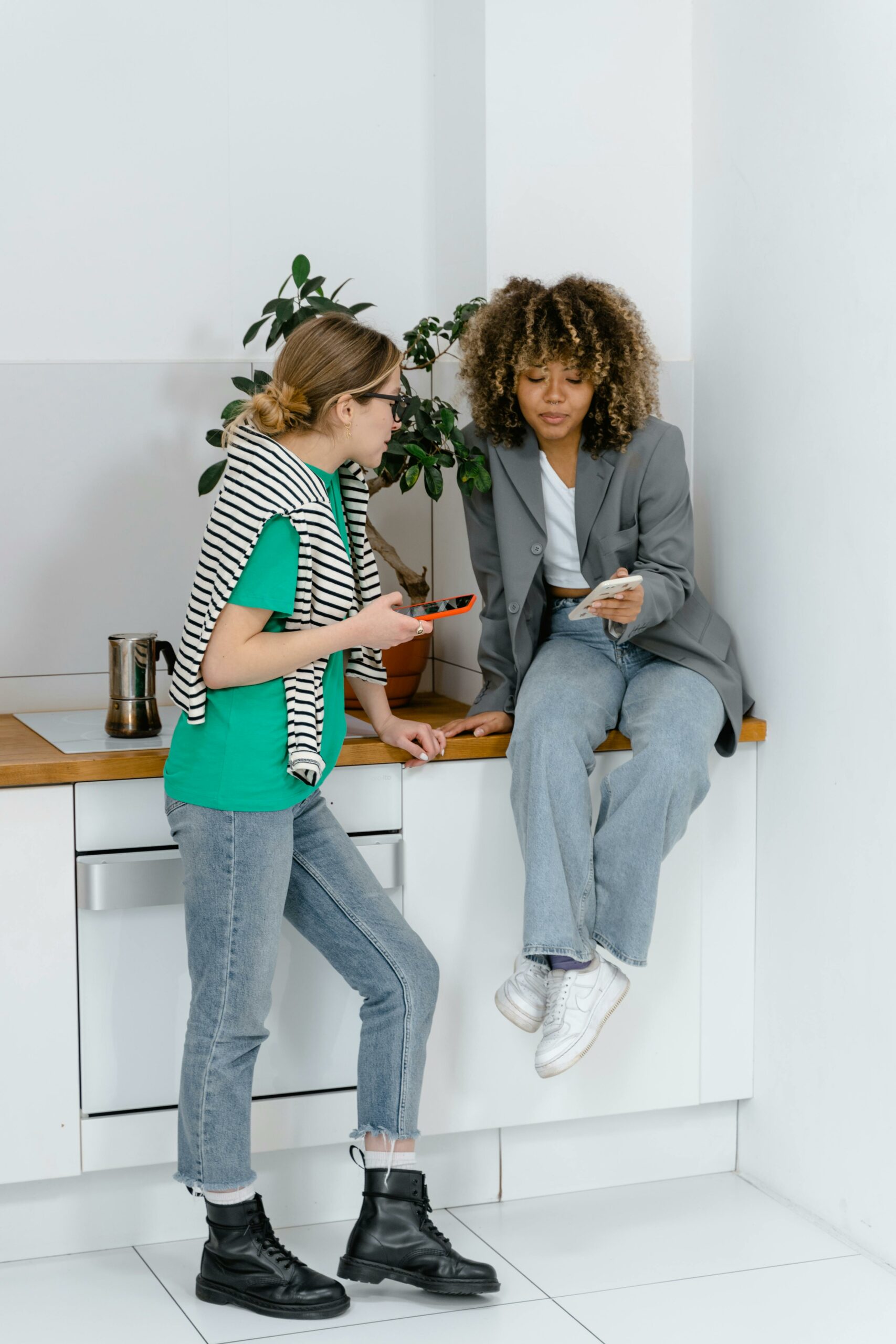 Two women casually chatting in a modern office wearing casual attire, using smartphones.